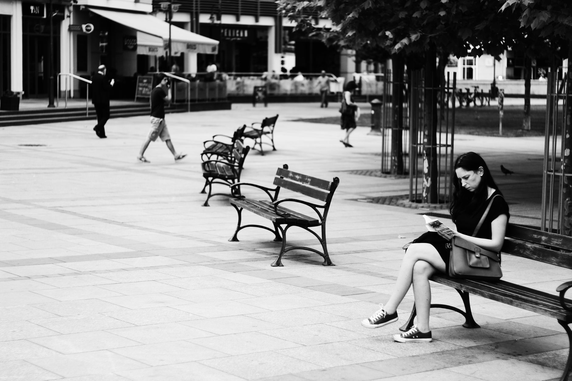 A woman in sneakers reads on a bench in a monochrome urban park setting.