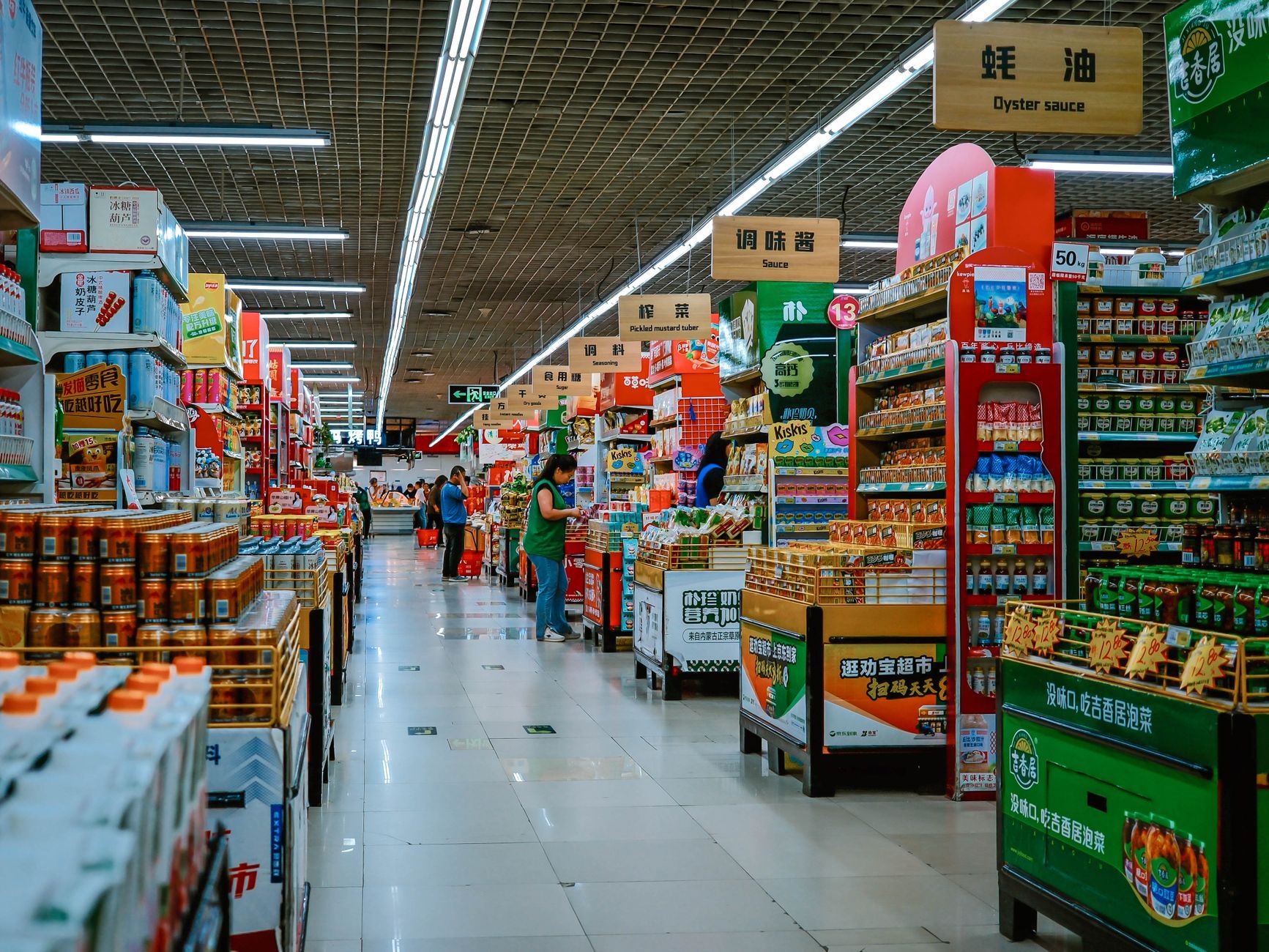 A active supermarket aisle filled with diverse products and shoppers under bright lighting.